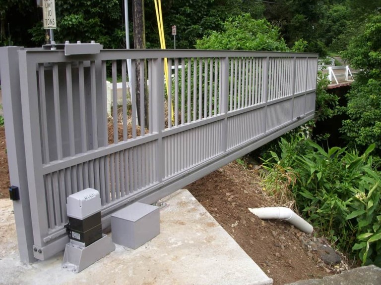 The sliding gate is made of gray metal, featuring vertical slats and is motorized, moving along a ground track for support.