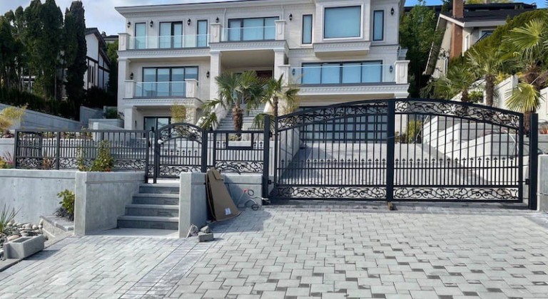 Elegant swing iron gates with intricate designs and pointed tops, installed at a residential driveway with metal fencing and concrete surroundings.