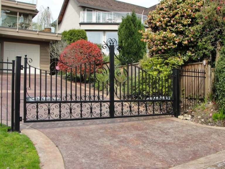 Decorative swing iron gates with ornate scrollwork, installed at a residential driveway, framed by lush landscaping.