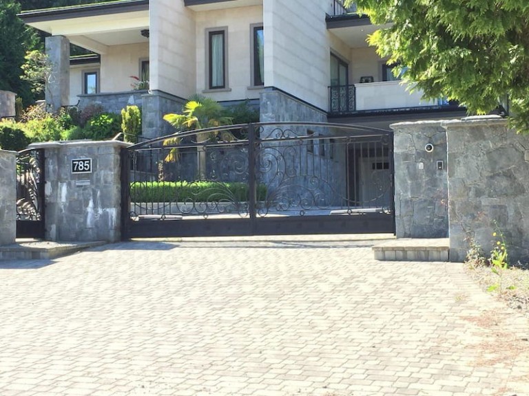 Elegant iron swing gate with decorative scrollwork, installed at a residential driveway between stone pillars.