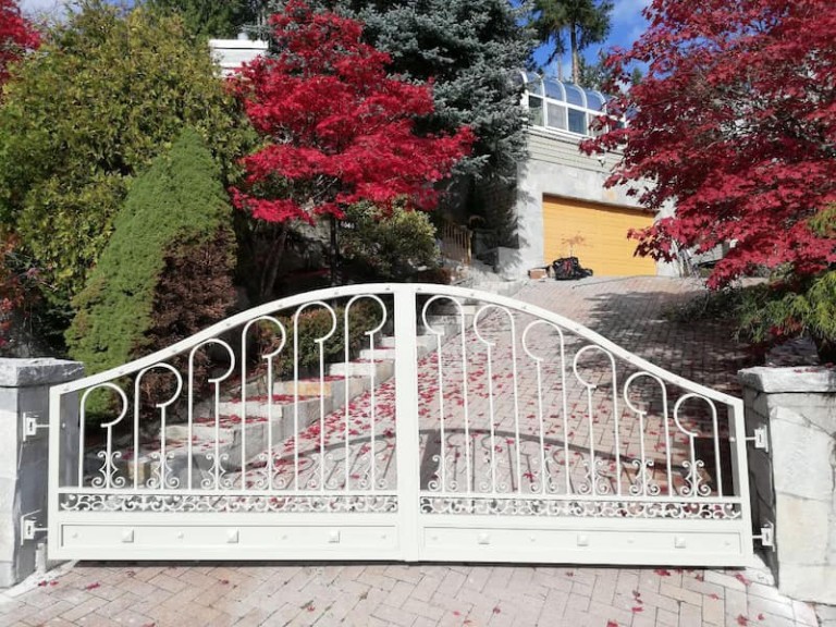 White iron swing gate with decorative patterns, installed at a residential driveway framed by stone pillars and vibrant landscaping.