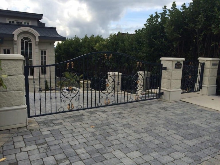 Ornate iron swing gate with gold accents, installed at a residential driveway, framed by stone pillars.