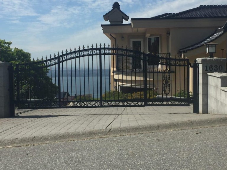 Black iron swing gate with pointed tops and decorative accents, installed at a residential driveway with stone pillars.