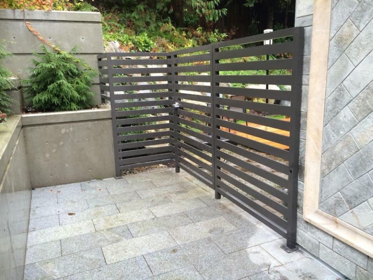 Metal pedestrian gate with horizontal slats, installed in a corner area with matching fencing and concrete surroundings.