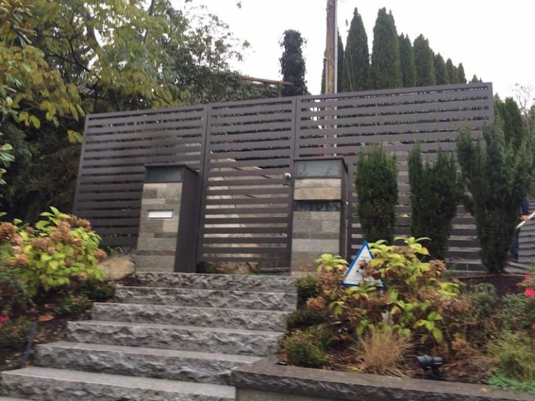 Modern pedestrian gate with horizontal metal slats, flanked by matching fencing and stone pillars, located at the top of a landscaped stone staircase.