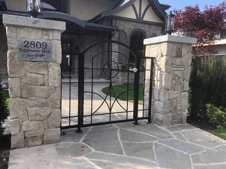 Elegant black pedestrian gate with a geometric design, framed by two stone pillars at the entrance of a residential pathway.